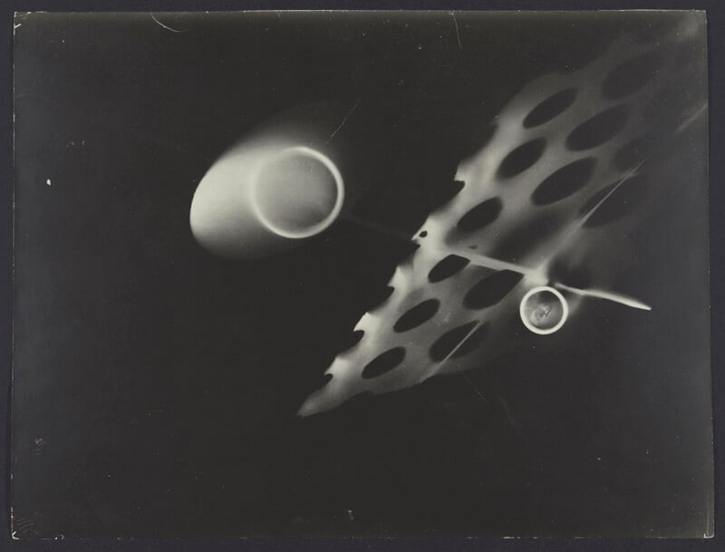 Abstract black and white photo featuring a translucent, perforated leaf-like shape, two circular forms, and soft, blurred light shapes on a dark background.