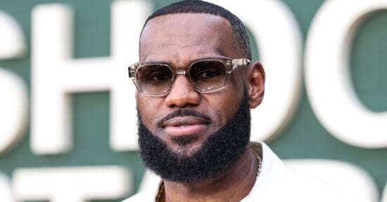 A man with a neatly groomed beard and mustache, wearing stylish sunglasses and a white shirt, stands in front of a green background with large, blurred white letters.