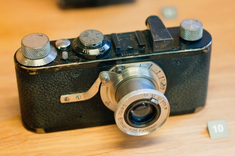 A vintage Leica camera with a 50mm lens sits on a wooden surface. The camera shows signs of wear, with scratches and worn edges, and there is a small numbered marker "10" nearby.
