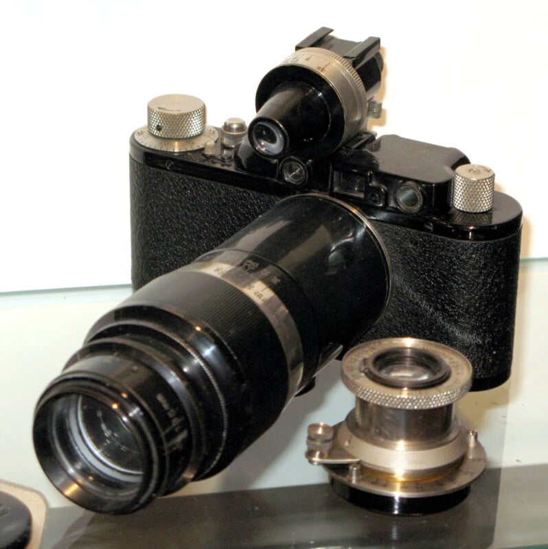 A vintage black camera with a large telephoto lens and an optical viewfinder, displayed on a clear surface with an additional lens attachment beside it.