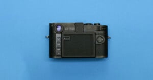A black digital camera with a textured grip is shown with its back facing up on a blue background, displaying control buttons, a viewfinder, and a display screen.