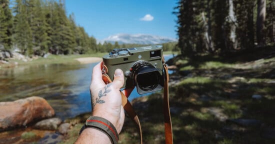 A tattooed hand holds a camera over a scenic outdoor landscape with a river, rocks, green trees, and distant mountains under a bright blue sky.