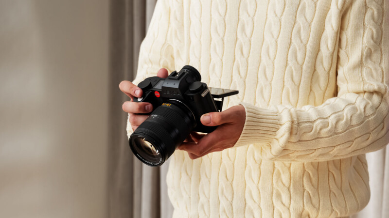 A person wearing a cream-colored cable knit sweater holds a black digital camera with a large lens and flip-out screen, standing indoors near a window with beige curtains.