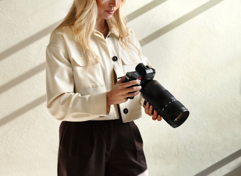 A woman with long blonde hair, wearing a cream-colored jacket and dark pants, holds a professional camera with a large lens, standing in soft natural light.