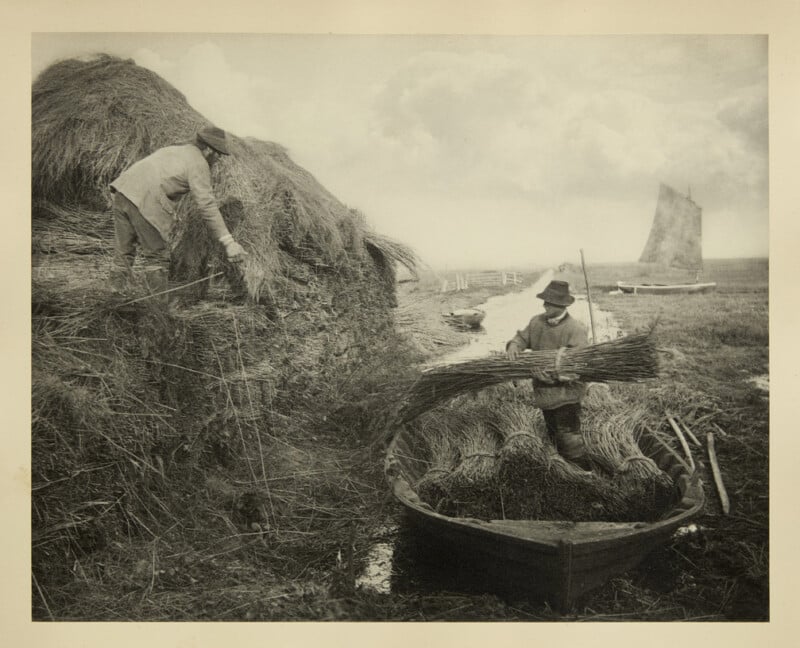 Two people gather and load bundles of reeds onto a small boat beside a large haystack in a marshy landscape. A sailboat is visible in the background under a cloudy sky.