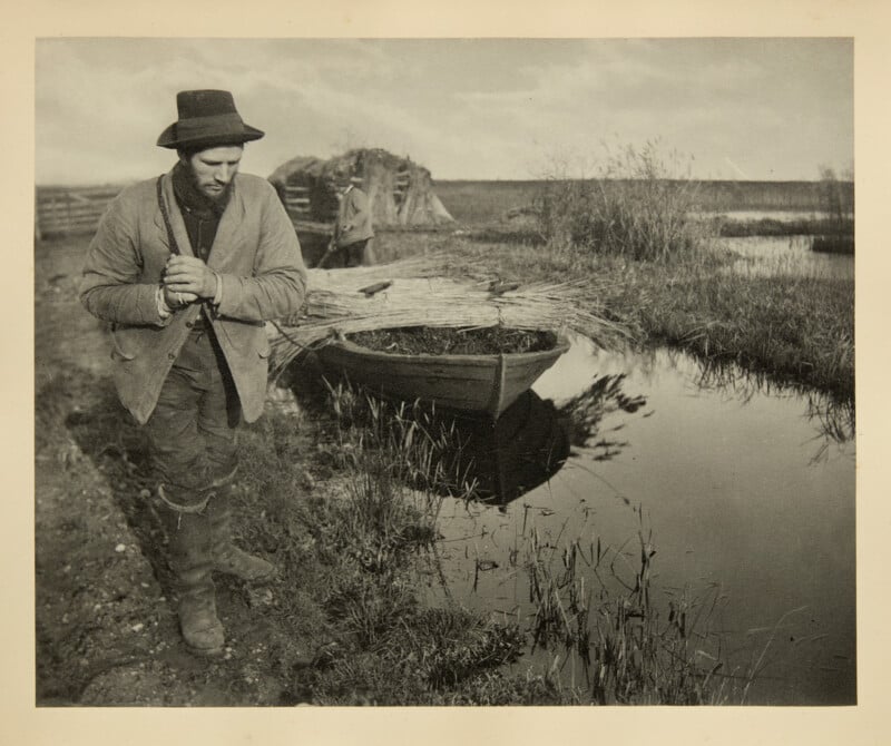 A man in rustic clothing and a hat stands by a small river, hands clasped. Behind him is a rowboat filled with reeds, grassy banks, reeds, and a wooden fence. The scene looks rural and tranquil.