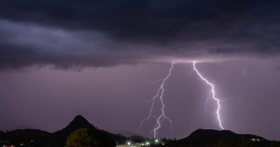 Several bolts of lightning illuminate a dark, cloudy night sky above distant hills and a small town, with silhouettes of trees visible in the foreground.