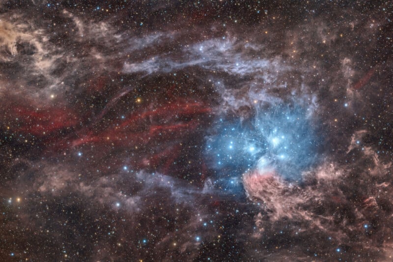 A colorful image of outer space features a star cluster glowing blue amid swirling clouds of red, pink, and gray nebulae, set against a background densely filled with stars.