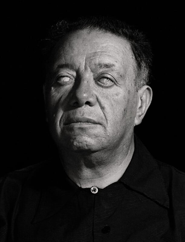 A black and white portrait of a middle-aged man with short, dark hair, wearing a dark shirt. He gazes slightly upward, and his eyes appear clouded or unfocused. The background is completely black.