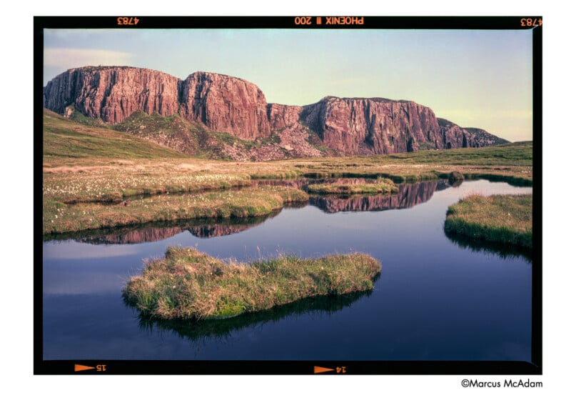 Rocky cliffs under a blue sky are reflected in a calm pond surrounded by grassy patches and fields, creating a serene natural landscape. The image has a classic film border and is credited to Marcus McAdam.