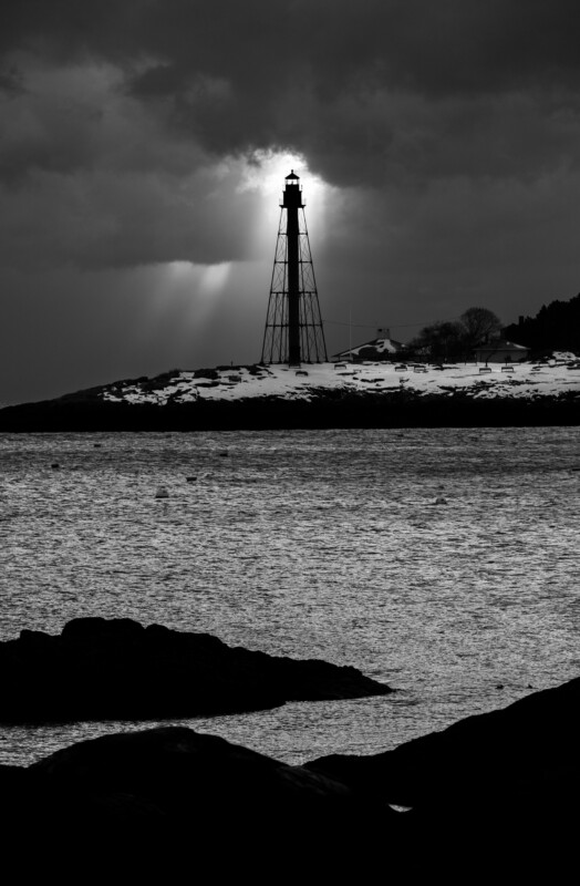 A tall lighthouse stands on a snowy, rocky shoreline under a cloudy sky. Sunbeams break through the clouds, illuminating the lighthouse as dark water and rocks fill the foreground. The image is in black and white.