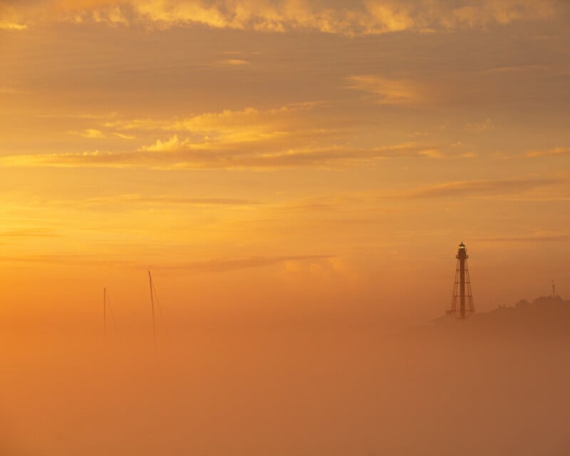 A lighthouse and sailboat masts partially obscured by thick orange mist at sunrise or sunset, with golden clouds filling the sky.