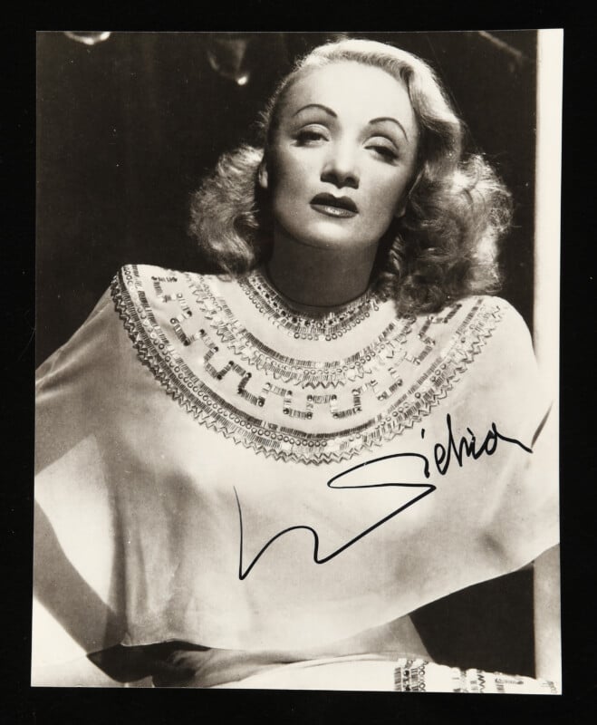 Black-and-white portrait of a woman with wavy hair, wearing an ornate, beaded top. She gazes upward with a serious expression. There is a large autograph signature across the lower part of the image.