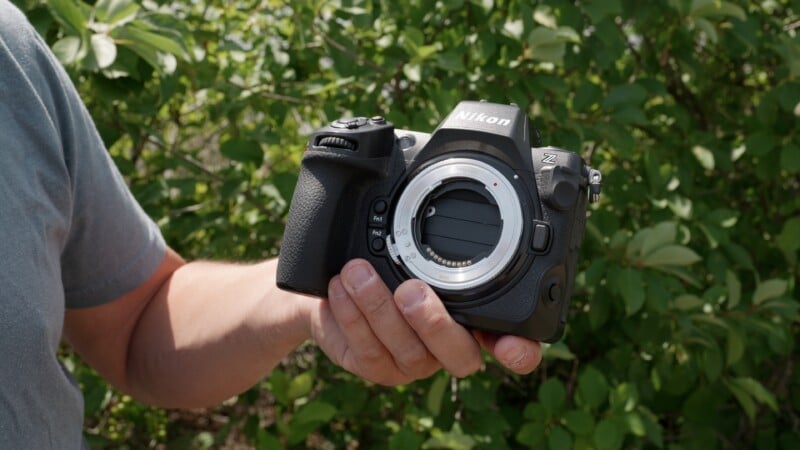 A person holding a Nikon Z series mirrorless camera body outdoors, with green leafy plants in the background. The camera does not have a lens attached.