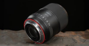A Meike 85mm f/1.8 camera lens is shown resting on a textured gray surface, with the focus on the metal lens mount and aperture markings. The background is black, highlighting the lens details.