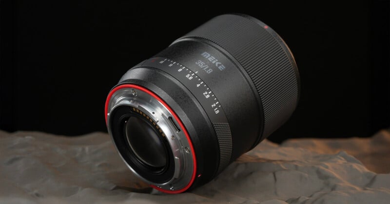 A Meike 85mm f/1.8 camera lens is shown resting on a textured gray surface, with the focus on the metal lens mount and aperture markings. The background is black, highlighting the lens details.