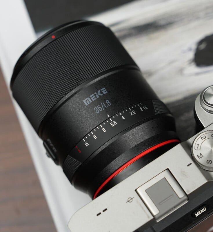 Close-up of a Meike 35mm f/1.8 camera lens attached to a silver mirrorless camera, with a blurred background and part of a textured surface visible.