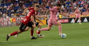 A soccer player in a pink uniform dribbles the ball past two opponents in red and black jerseys during a match, with a blurred crowd in the background.