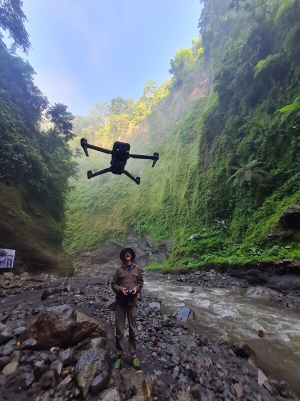 A person operating a drone stands on rocky ground beside a flowing river, surrounded by lush green cliffs and dense vegetation under a bright, clear sky.