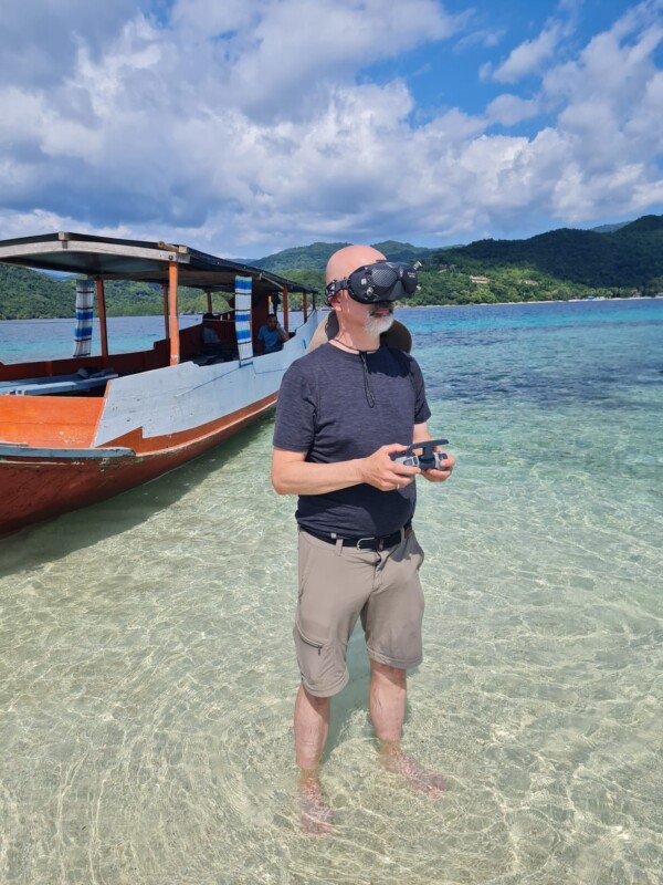A man stands knee-deep in clear, shallow water near a wooden boat, wearing VR goggles and holding a remote control. Behind him are green hills and a partly cloudy sky.