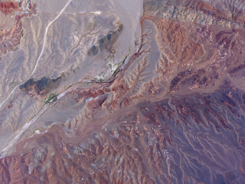 Aerial view of an arid, mountainous landscape with rugged, reddish-brown terrain, winding valleys, and sparse patches of green vegetation. Pale flatlands and a light road run across the upper left corner.