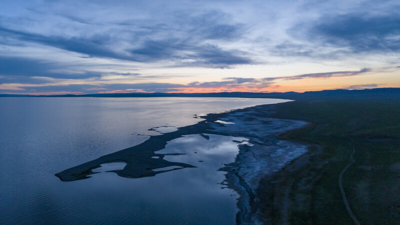 A serene aerial view of a lake at sunset, with calm water reflecting the colorful sky. The shoreline curves gently, bordered by patches of land and a dark silhouette of distant hills.