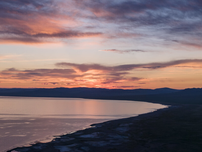 A serene sunset over a calm body of water, with orange, pink, and purple hues in the sky, reflecting on the surface. Dark silhouettes of distant hills and shoreline are visible under the colorful clouds.