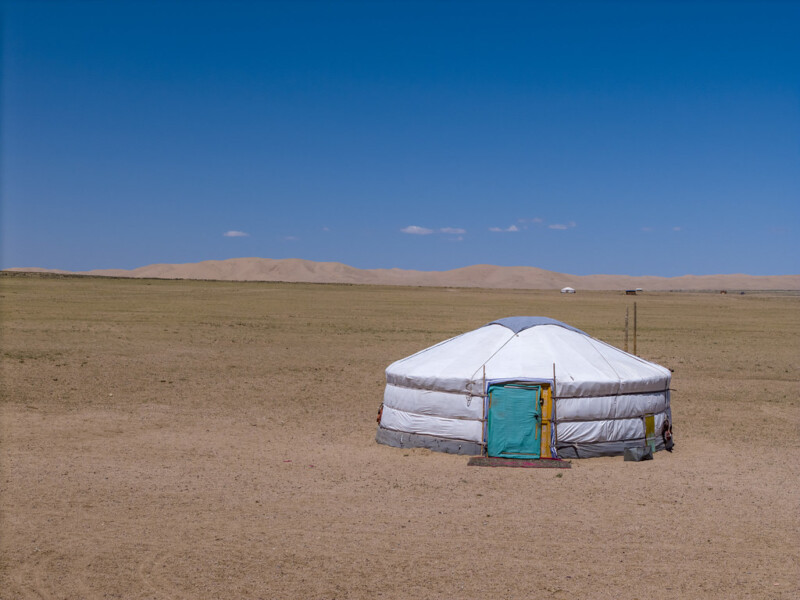 A white traditional yurt with a blue door stands alone on a vast, dry plain under a clear blue sky, with distant hills visible in the background.