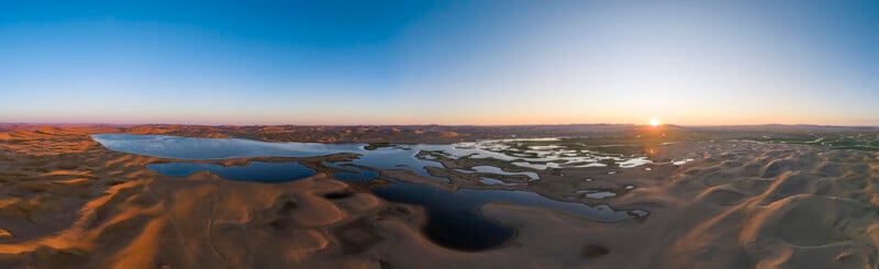 A panoramic aerial view of a desert landscape with sand dunes, scattered water pools, and a lake, captured at sunrise under a clear blue sky.