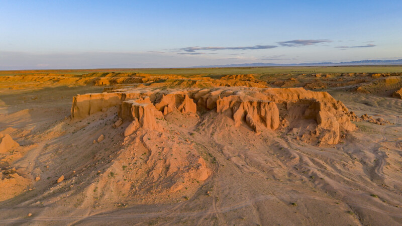 Aerial view of the Flaming Cliffs in Mongolia, showing orange sandstone formations under a clear blue sky with sunlight casting shadows on the arid landscape.