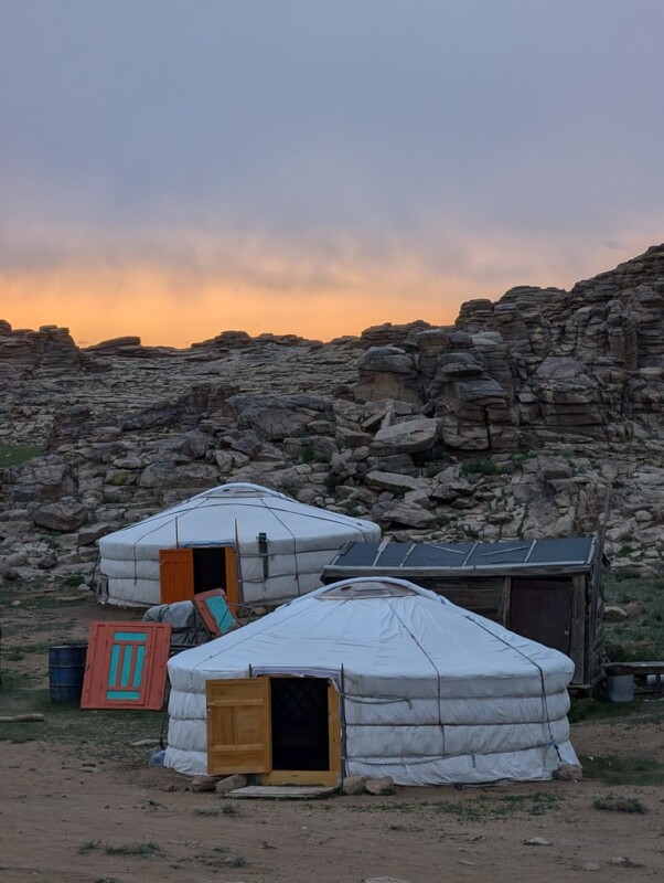 Two traditional white yurts with colorful doors stand on a rocky landscape at sunset, with orange light illuminating the sky and rugged cliffs in the background.