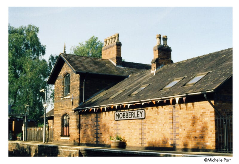 A brick railway station building with chimneys and a sign reading "Mobberley." Bunting is strung along the roof, and trees are visible in the background under a clear sky.