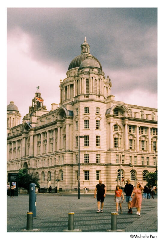 A grand, historic building with a domed roof stands under a cloudy sky. Several people walk in the foreground, and the architecture features tall windows and ornate details.