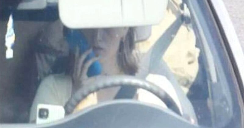 A person is driving a car while holding a phone to their ear, partially obscured by the rearview mirror and steering wheel. The person appears distracted while operating the vehicle.