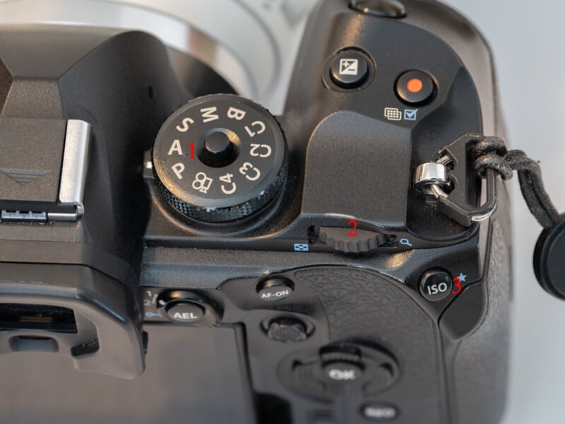 Close-up of an OM System OM-1 digital camera's top controls, showing the mode dial with various shooting options, shutter button, ISO button, and part of the camera strap attachment.
