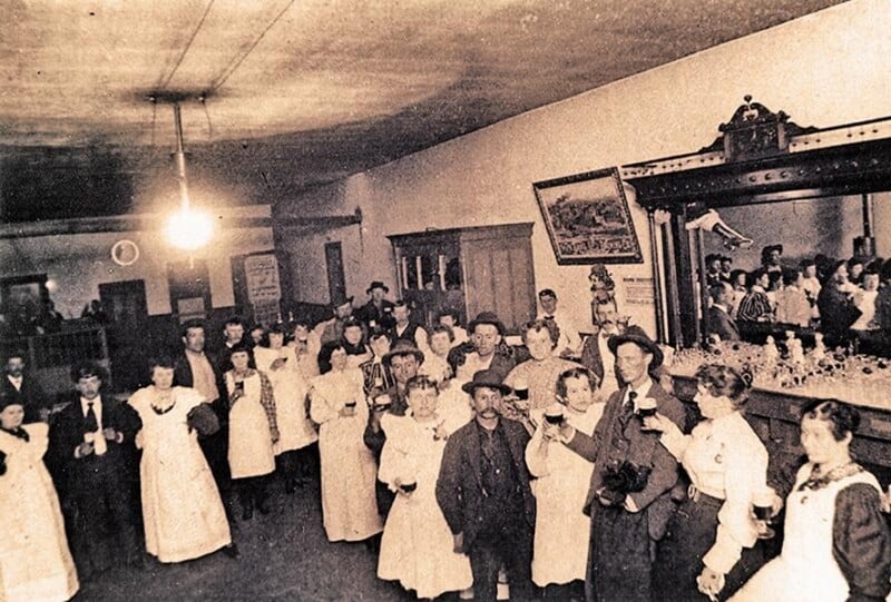 A sepia-toned historical photo of men and women gathered in a bar or saloon, many holding drinks, with a large mirror behind the bar and a glowing ceiling light overhead. The scene appears lively and social.