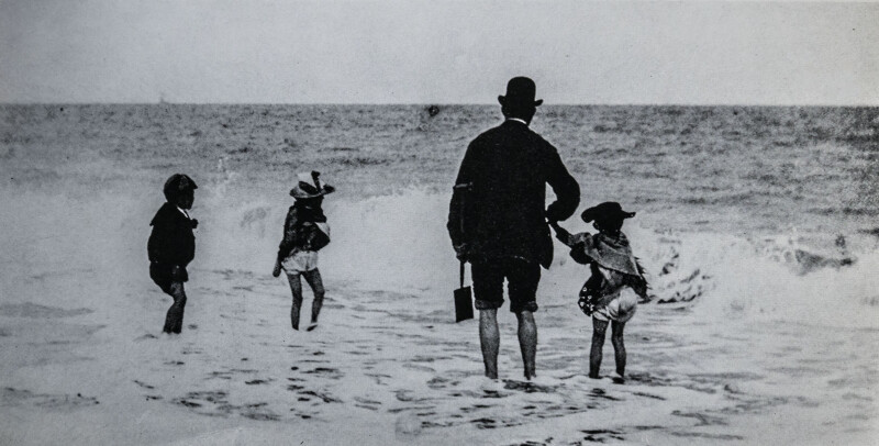 A man and three children stand at the edge of the sea, with their backs to the camera, playing in shallow waves on a beach under an overcast sky.