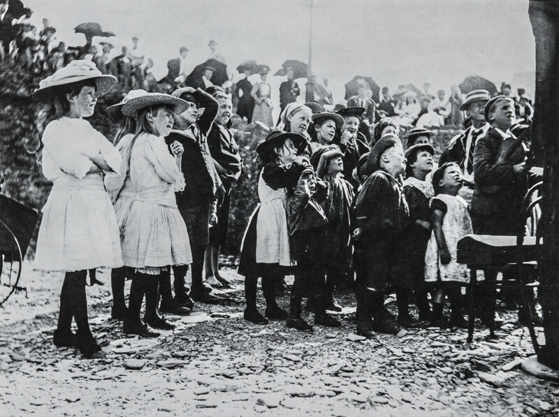 A group of children in early 20th-century clothing gather outdoors, looking up with interest at someone or something out of frame; a crowd of adults stands behind them, some holding umbrellas.