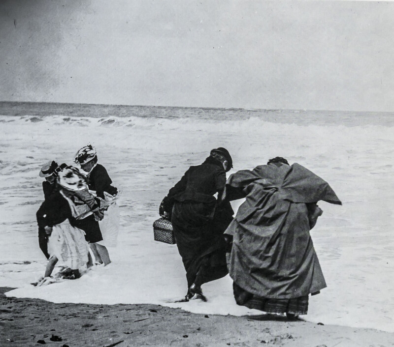 Four women in old-fashioned clothing stand at the edge of the sea, waves touching their feet. Two hold hands, and one carries a basket. The scene appears breezy and playful on a sandy beach.
