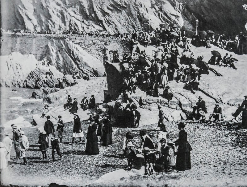 A large group of people, dressed in late 19th or early 20th-century clothing, gather on a rocky beach and nearby cliffs, some sitting and some standing, with a backdrop of rugged rocks.