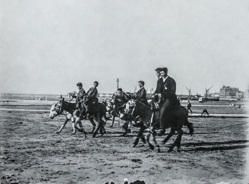 A group of people ride ponies along a sandy beach. In the background, there are a few people walking, ships, and distant buildings near the shoreline under a clear sky.