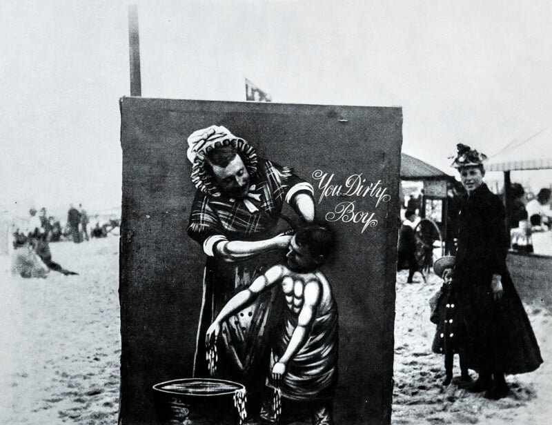 A vintage black-and-white photo shows a beach scene with a painted cutout of a woman washing a boy, labeled "You Dirty Boy," with a woman standing nearby and people in the background.