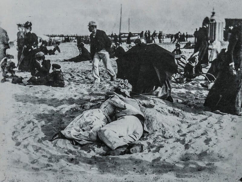 Historic black and white photo of people relaxing on a crowded beach; a couple lies close together in the sand in the foreground, while groups of people stand, walk, and sit in the background.