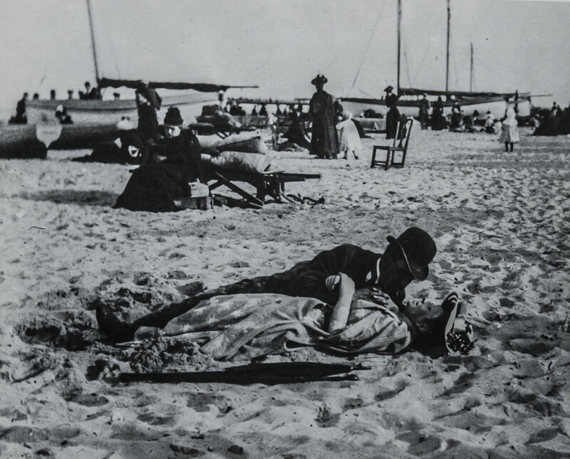 A vintage black-and-white photo shows a couple lying on a sandy beach, embracing and kissing. People sit and walk in the background near boats, with some dressed in early 20th-century clothing.