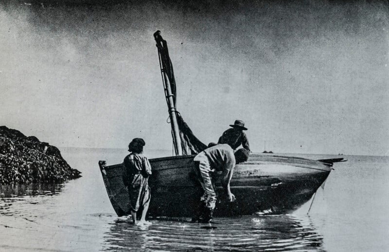 A black and white photo of three people beside a small wooden boat at the shore, with shallow water around their feet. One person bends toward the boat, another stands nearby, and a child stands to the left.
