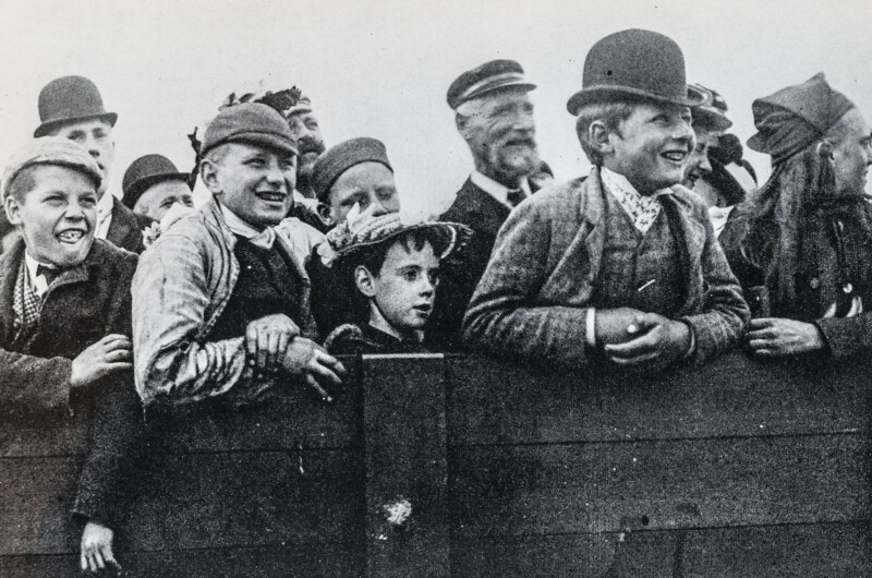 A group of boys and men in old-fashioned clothing smile and lean over a wooden fence, watching something with excitement and interest; the photo is in black and white.