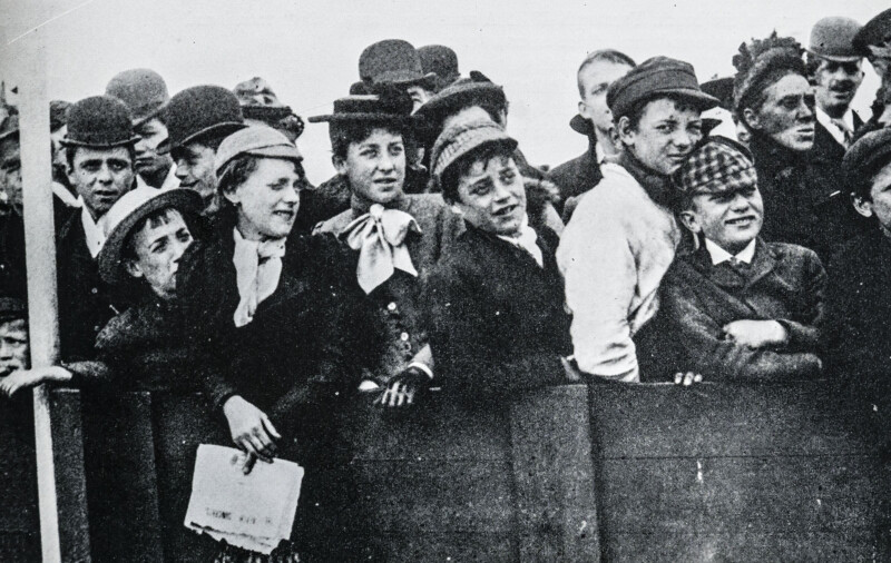 A black-and-white photo of a group of young people and men, some wearing caps and hats, smiling and leaning over a wooden barrier while watching an event. The scene appears to be from the early 20th century.