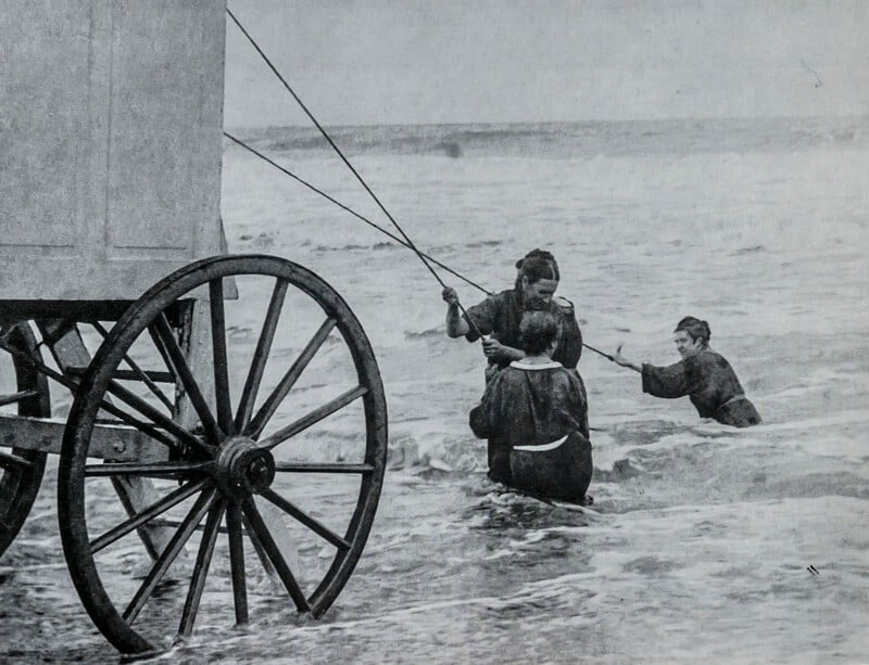 Three women in old-fashioned swimsuits hold onto ropes attached to a wheeled bathing machine as they stand and kneel in the surf, with waves crashing around them at the beach.