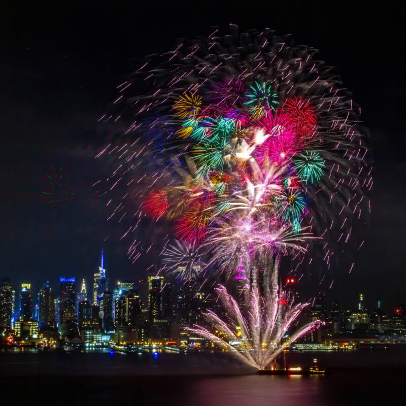 Colorful fireworks explode in the night sky above a city skyline, with tall buildings illuminated and their reflections visible on the water below.