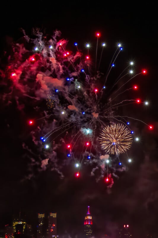 Red, white, and blue fireworks burst in the night sky above a city skyline, with the Empire State Building illuminated in matching colors below. Smoke from the fireworks partially obscures some city lights.